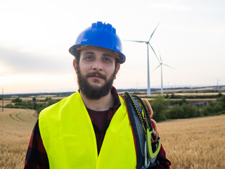 Smiling worker man with safety helmet and work clothes in a field of windmills looking at camera. Renewable energies, green energies 