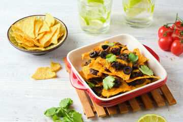 Hot appetizer, baked corn chips with refried beans, cheddar cheese and olives in a ceramic form on a light wooden background. Appetizer recipes. Party, fast food.