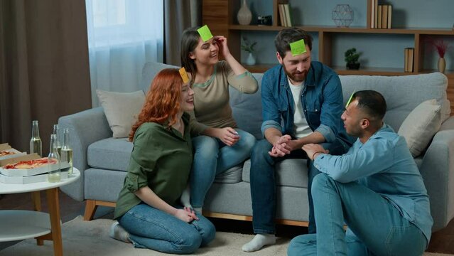 Four diverse friends play guessing game with sticky notes on head creative multiracial women men in living room playing intellectual charades asking questions Who am I talking laughing funny playtime