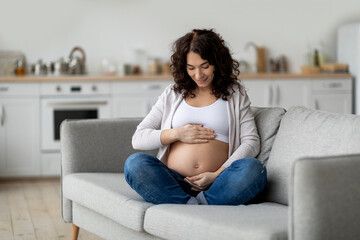 Happy Pregnant Woman Tenderly Embracing Belly While Sitting On Couch At Home