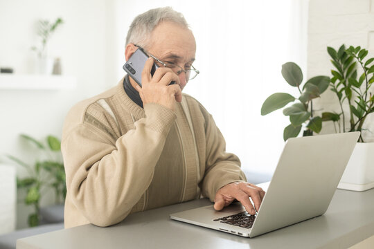 Handsome smiling senior man wearing glasses using mobile phone while sitting at his cozy workplace with laptop at home, retired male chatting with friends in social media, typing on smartphone.