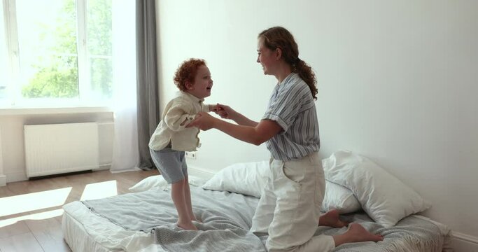 Cheerful woman plays with adorable toddler 2s redhead son, holding hands, jumping on bed, enjoy funny lively active game in warm cozy sunlit bedroom. Carefree, happy pastime of mother and little child