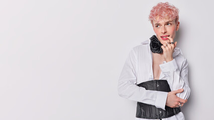 Androgynous man with pensive expression bites finger focused aside wears white shirt and black...
