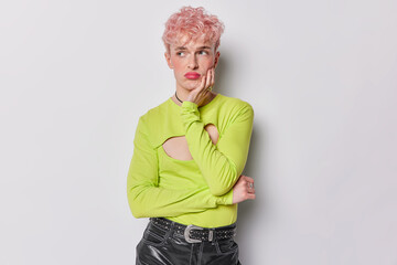 Horizontal shot of displeased thoughtful gay with makeup dressed in women clothes keeps hand on cheek looks thoughtful and dissatisfied aside isolated over white background. Transgender man indoors