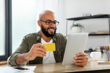 Online shopping. Excited latin businessman using tablet and credit card, making payment via bank app, sitting at desk