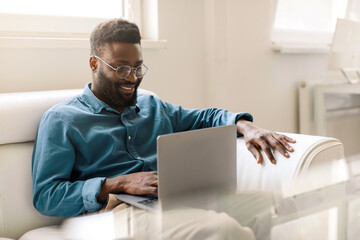 Happy african american businessman sitting in office on couch, working on laptop computer and...