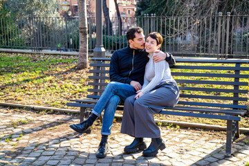 The young couple is sitting on a bench in the park in Rome. Beautiful couple kisses in the park. - 575648332