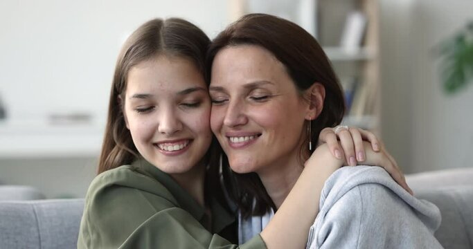 Woman sits on sofa with teen daughter, hug, smile look at cam, enjoy moment of tenderness. Close up happy faces two generations women, feel affection, having strong family ties, celebrate Mothers Day