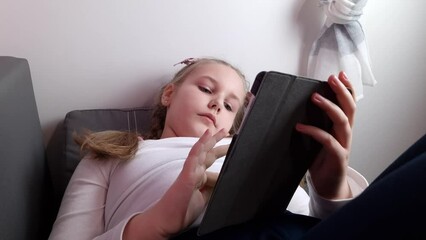 Serious little caucasian child girl use tablet computer, play online game lying on bed. Modern technology, childhood concept