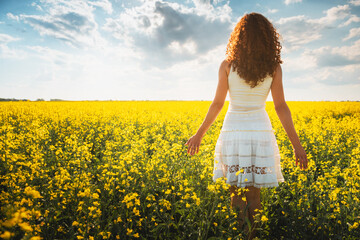 Woman with long curly hair stands in a field with yellow flowers and enjoying the sunset