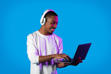 Joyful black guy using modern laptop and wireless headphones, gaming