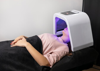LED light mask for facial skin therapy, care. Young woman gets skin rejuvenation light treatment, lying on couch under blue mask. Stimulation of collagen production, killing acne bacteria. Horizontal.