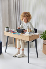 Vertical shot of curly haired woman draws sketches on graphic tablet uses stylus creats interior design for apartment plans repairment wears striped jumper and slippers poses over cozy home interior