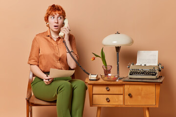 Shocked redhead adult woman talks via stationary phone has amazed expression holds paper works in own cabinet with old furniture wears shirt and green trousers isolated over brown background