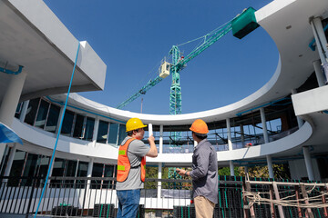 Two young structural engineer and architect dressed in orange work vests standing in front of mechanical crane working on the construction site.