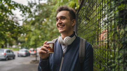 One young adult caucasian man in the city or town near park with cup of coffee in autumn or spring day happy male tourist standing alone wait real people copy space