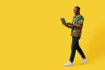Joyful black man in african costume using laptop on yellow
