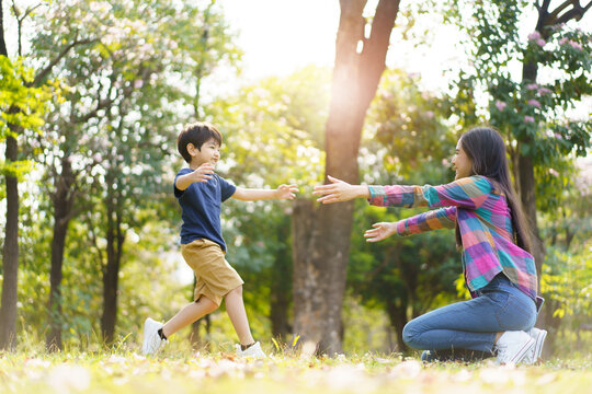 Asian little cute boy running to hug his mother at the park, mother and sun relaxation activity in a park or garden in weekend. Woman and a boy playing on a beautiful sunlight and bokeh background.