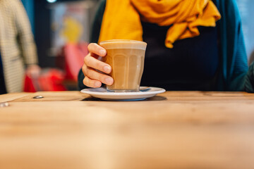 Woman holding coffee cup with frothy coffee while wearing yellow scarf, siting at wooden table in cafe