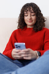 Beautiful white girl sitting on couch and typing a message on a smart phone. Cute female person with curly hair using modern blue mobile phone