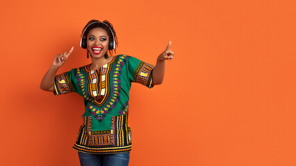 Positive black lady in african costume using wireless headphones