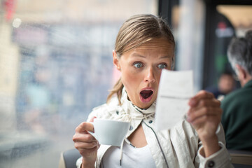 Pretty, young woman baffled with the bill in a coffeeshop/restaurant - obrazy, fototapety, plakaty