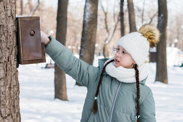 A teenage girl pours seeds into a bird feeder on a tree in a winter forest. Walk outdoors.