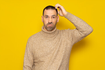 Doubtful Hispanic man thinking, scratching his head trying to find a solution, isolated on a yellow background. He tries to decide where to spend the holidays.