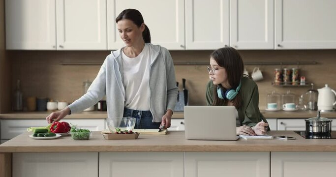 Family spend weekend at home, middle-aged mom prepare vegetarian salad, cuts fresh celery for healthy dish and talks to teen daughter, makes notes, studying with computer use modern tech in kitchen