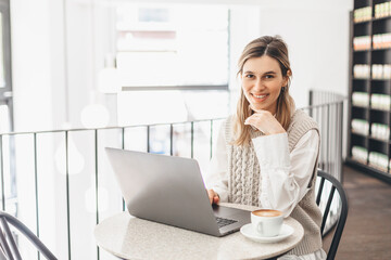 Image of young beautiful joyful woman smiling while working with laptop in office or cafe with coffee on table. Woman wear knitted vest and white shirt, hold her head. Good work day.