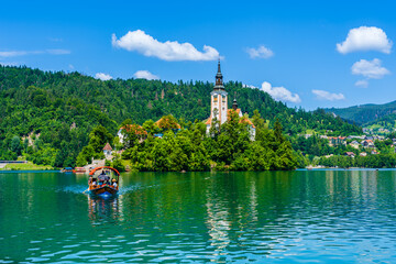 Fototapeta na wymiar Bled, Slovenia: Sailing boat towards Bled island and church on lake Bled; reflection on the water surface