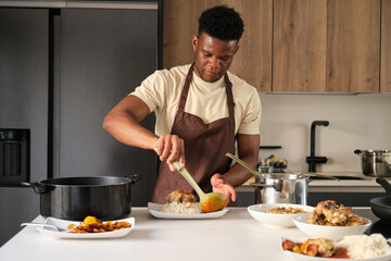 Fototapeta na wymiar Young african man serving tomato sauce on fried chicken mince and rice on a plate in a kitchen.