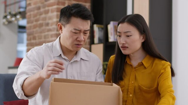Couple of asian spouses opening delivery box, looking inside and feeling displeased, disappointed with bad purchase