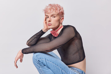 Non-binary person keeps hand on face looks directly at camera squats against white background wears...