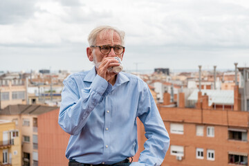Mature businessman with gray hair and beard in blue shirt, jeans and glasses drink coffee on rooftop terrace with city skylight on background. Copy space