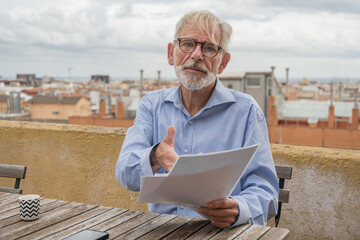 Mature businessman with gray hair and beard in blue shirt, jeans and glasses with paper documents on rooftop terrace with city skylight on background. Copy space