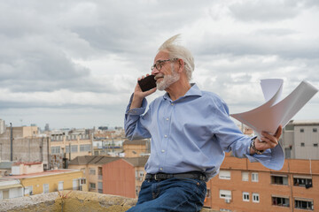 Mature businessman with gray hair and beard in blue shirt, jeans and glasses with paper documents and smartphone on rooftop terrace with city skylight on background. Copy space