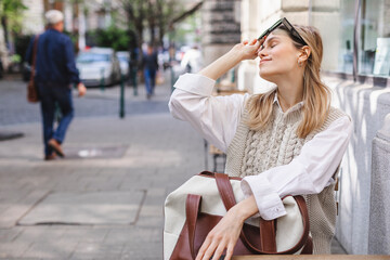 Fototapeta na wymiar Young woman sitting on a street cafe waiting order, holding bag in arms and watching the city life, enjoy Europe lifestyle. Girl take off black sunglasses, wear white shirt and beige knitted vest, bag