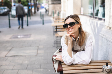 Fototapeta premium Young woman sitting on a street outside in coffee shop waiting order, holding her head and look at camera.