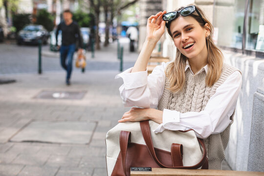 Young woman sitting on a street cafe waiting order, holding bag in arms and watching the city life, enjoy Europe lifestyle. Girl take off black sunglasses, wear white shirt and beige knitted vest, bag