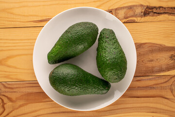 Three juicy avocados with a white ceramic plate on a wooden table, macro, top view.