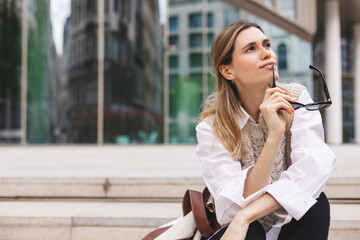 Fototapeta premium Beautiful smiling fashionable businesswoman sitting on the stairs in front of business center, she look at side, dreaming or thinking, bite shackle of sunglasses. Woman wear knit vest, white shirt.