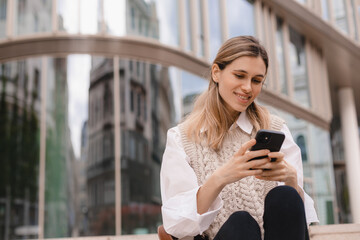Fototapeta premium Low angle of glad woman in formal wear sitting in megapolis on street near urban building using smartphone, look happy and smiling. Blonde girl read good news.