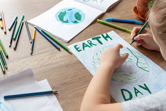 A Child At Home At The Table Draws The Planet Earth With A World Map With Multi-colored Pencils And Felt-tip Pens On White Paper. Peace And Earth Day Concept.