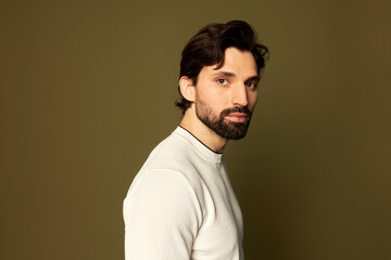 Picture of millennial manful guy in white casual clothes posing against brown studio background, turned by his name, looking at camera, having stylish haircut and beard. Handsome male appearance