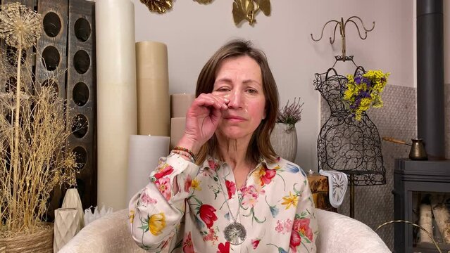 Sad middle-aged woman in casual clothing sharing negative emotions indoors. Unhappy adult female expressing distress, pain sorrow, crying, wiping tears off with hand sitting on couch at interior room