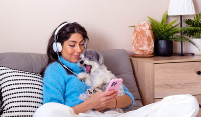 Young woman with headphones sitting at home with her dog listening ebook with her mobile phone 