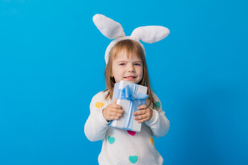 a happy little girl with bunny ears on her head and a gift box on a blue background. Easter concept, space for text