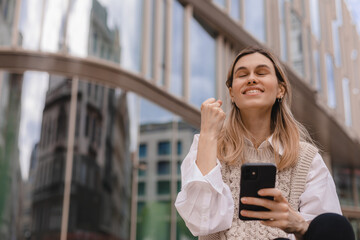 Beautiful smiling fashionable businesswoman sitting on the stairs in front of business center and show winner gesture, hold mobile phone, look happy. Amazed woman wear beige knit vest, white shirt.