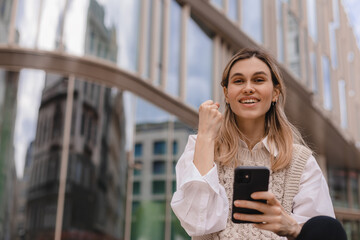 Beautiful smiling fashionable businesswoman sitting on the stairs in front of business center and show winner gesture, hold mobile phone, look happy. Amazed woman wear beige knit vest, white shirt.